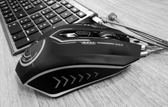 A sleek, futuristic-looking computer mouse with intricate design patterns sits on a wooden desk next to a keyboard. The mouse features smooth lines and geometric shapes, and the keyboard keys are clearly visible.