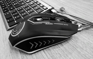A sleek wireless mouse resting on a wooden desk beside a glowing keyboard.