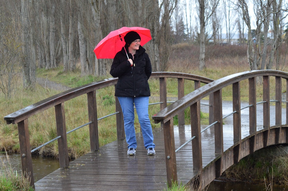 woman in black jacket and blue denim jeans holding red umbrella walking on wooden bridge during