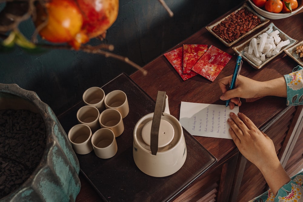 person writing on white paper beside white ceramic mug