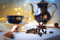 A cozy table setting featuring a star anise and peppercorns on a tablecloth, accompanied by a spoon filled with spice. In the background, a blurred teapot and teacup are illuminated by soft, warm bokeh lights, creating a serene and inviting atmosphere.