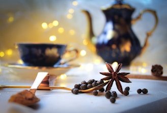 A cozy table setting featuring a star anise and peppercorns on a tablecloth, accompanied by a spoon filled with spice. In the background, a blurred teapot and teacup are illuminated by soft, warm bokeh lights, creating a serene and inviting atmosphere.