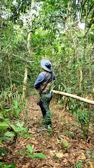 A person wearing a wide-brimmed hat and vest stands in a dense, lush rainforest environment, surrounded by vibrant greenery, vines, and a variety of plants. They appear to be exploring or observing the natural surroundings.
