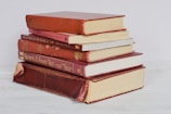 A stack of six hardcover books, mostly in shades of red, is arranged neatly on a light-colored surface. Some of the books have visible wear, with faded covers and slightly frayed edges.