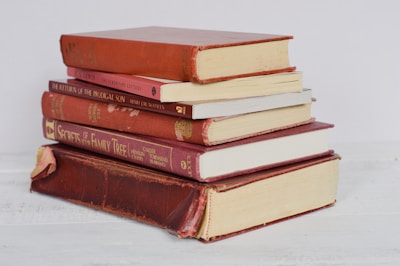 A stack of six hardcover books, mostly in shades of red, is arranged neatly on a light-colored surface. Some of the books have visible wear, with faded covers and slightly frayed edges.