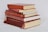 A stack of six hardcover books, mostly in shades of red, is arranged neatly on a light-colored surface. Some of the books have visible wear, with faded covers and slightly frayed edges.