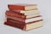 A stack of six hardcover books, mostly in shades of red, is arranged neatly on a light-colored surface. Some of the books have visible wear, with faded covers and slightly frayed edges.