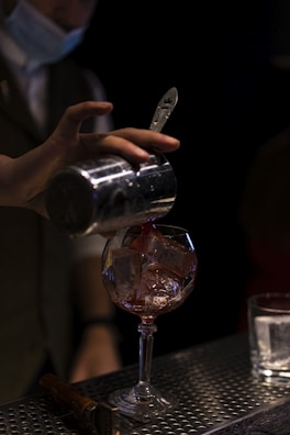 A bartender wearing a face mask is pouring a red liquid from a metal shaker into a glass filled with ice cubes. The setting appears to be a dimly lit bar or lounge area. On the counter, there is another clear glass with ice and a bar tool, possibly a muddler or a bottle opener.