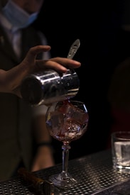 A bartender wearing a face mask is pouring a red liquid from a metal shaker into a glass filled with ice cubes. The setting appears to be a dimly lit bar or lounge area. On the counter, there is another clear glass with ice and a bar tool, possibly a muddler or a bottle opener.