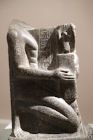 A stone sculpture depicting a partially damaged seated human figure with intricate hieroglyphic carvings. The figure holds an object close to its chest, and the overall texture of the statue is rough with visible wear, indicating its age. The artwork is positioned against a plain background, highlighting its detailed craftsmanship.