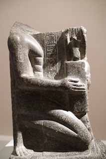 A stone sculpture depicting a partially damaged seated human figure with intricate hieroglyphic carvings. The figure holds an object close to its chest, and the overall texture of the statue is rough with visible wear, indicating its age. The artwork is positioned against a plain background, highlighting its detailed craftsmanship.