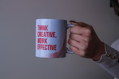 A hand holding a white mug with red text that reads 'THINK CREATIVE, WORK EFFECTIVE.' The background is plain and neutral, and the person is wearing a light-colored sweater with button details on the cuff.