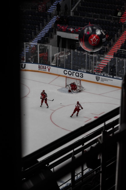 An ice hockey game with players in red uniforms on the rink, a goalie in front of the net, and a large inflatable with a star logo floating above. The arena has tiered seating with illuminated stairs, and various banners and signs around the rink.
