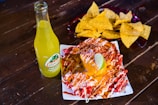 A vibrant scene with a bottle of Jarritos pineapple soda next to a plate of spicy snacks covered in melted cheese and spices, garnished with a lime wedge. In the background, a bowl filled with tortilla chips is visible, all placed on a rustic wooden table.