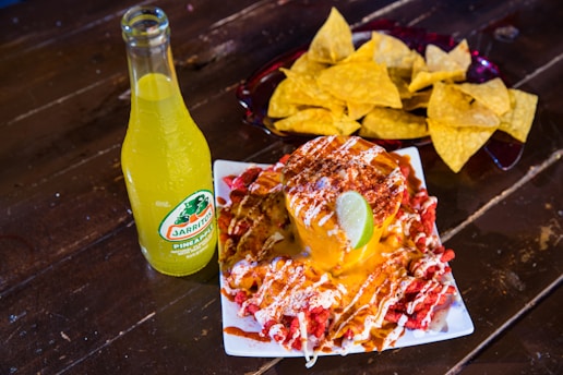 A vibrant scene with a bottle of Jarritos pineapple soda next to a plate of spicy snacks covered in melted cheese and spices, garnished with a lime wedge. In the background, a bowl filled with tortilla chips is visible, all placed on a rustic wooden table.