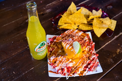 A vibrant scene with a bottle of Jarritos pineapple soda next to a plate of spicy snacks covered in melted cheese and spices, garnished with a lime wedge. In the background, a bowl filled with tortilla chips is visible, all placed on a rustic wooden table.