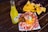 A vibrant scene with a bottle of Jarritos pineapple soda next to a plate of spicy snacks covered in melted cheese and spices, garnished with a lime wedge. In the background, a bowl filled with tortilla chips is visible, all placed on a rustic wooden table.