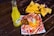 A vibrant scene with a bottle of Jarritos pineapple soda next to a plate of spicy snacks covered in melted cheese and spices, garnished with a lime wedge. In the background, a bowl filled with tortilla chips is visible, all placed on a rustic wooden table.