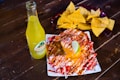 A vibrant scene with a bottle of Jarritos pineapple soda next to a plate of spicy snacks covered in melted cheese and spices, garnished with a lime wedge. In the background, a bowl filled with tortilla chips is visible, all placed on a rustic wooden table.