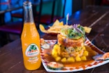 A bottle of Jarritos Mango soda is placed on a wooden table next to a plate of food. The food includes a stacked dish with various ingredients including avocado, greens, and a slice of red onion on top. The dish is garnished with a sauce and surrounded by pieces of cucumber. A basket of tortilla chips is visible in the background.