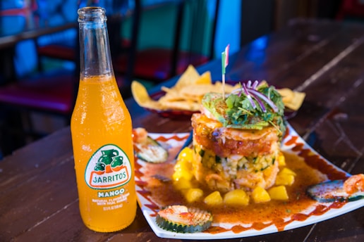 A bottle of Jarritos Mango soda is placed on a wooden table next to a plate of food. The food includes a stacked dish with various ingredients including avocado, greens, and a slice of red onion on top. The dish is garnished with a sauce and surrounded by pieces of cucumber. A basket of tortilla chips is visible in the background.