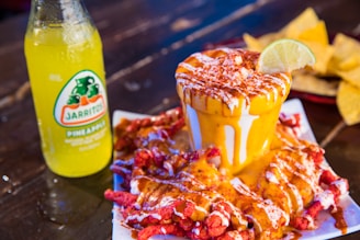 A cup filled with what appears to be cheese sauce, drizzled with chili powder and topped with a lime slice. Surrounding the cup are red spiced corn chips. To the side, there is a bottle of Jarritos pineapple soda, and in the background, some tortilla chips can be seen.