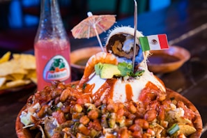 A vibrant plate filled with nachos topped with beans, diced vegetables, and drizzled with a red sauce. A scoop of ice cream with chocolate and coconut is placed on top, adorned with a small paper umbrella and a Mexican flag. A bottle of pink Jarritos soda stands in the background beside some tortilla chips.