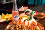 A vibrant Mexican dish is presented on a wooden table. It features a mound covered in a variety of toppings such as beans, avocado slices, and spicy sauce. Garnished with a small umbrella, the dish is stylishly topped with a rolled dessert resembling ice cream, as well as a tiny Mexican flag toothpick. Accompanying the food is a glass bottle of Jarritos soda with a pinkish hue and an arrangement of tortilla chips in the background.