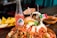 A vibrant Mexican dish is presented on a wooden table. It features a mound covered in a variety of toppings such as beans, avocado slices, and spicy sauce. Garnished with a small umbrella, the dish is stylishly topped with a rolled dessert resembling ice cream, as well as a tiny Mexican flag toothpick. Accompanying the food is a glass bottle of Jarritos soda with a pinkish hue and an arrangement of tortilla chips in the background.