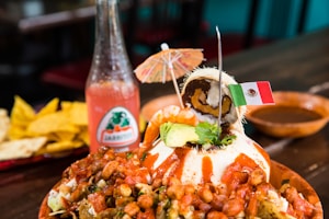 A vibrant Mexican dish is presented on a wooden table. It features a mound covered in a variety of toppings such as beans, avocado slices, and spicy sauce. Garnished with a small umbrella, the dish is stylishly topped with a rolled dessert resembling ice cream, as well as a tiny Mexican flag toothpick. Accompanying the food is a glass bottle of Jarritos soda with a pinkish hue and an arrangement of tortilla chips in the background.