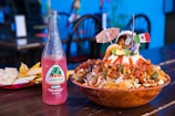 A vibrant table setting featuring a wooden bowl filled with a colorful Mexican dish topped with peanuts, avocado, and shrimp, garnished with a small festive umbrella and a Mexican flag. A pink Jarritos Guava soda bottle stands next to the bowl on a wooden table, accompanied by a plate of tortilla chips and quesadillas in the background. The setting is lively, suggesting a festive meal.
