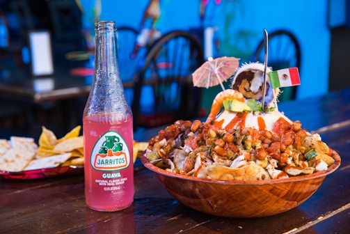A vibrant table setting featuring a wooden bowl filled with a colorful Mexican dish topped with peanuts, avocado, and shrimp, garnished with a small festive umbrella and a Mexican flag. A pink Jarritos Guava soda bottle stands next to the bowl on a wooden table, accompanied by a plate of tortilla chips and quesadillas in the background. The setting is lively, suggesting a festive meal.
