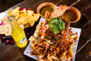 A vibrant Mexican dish featuring a large serving of shrimp cocktail topped with avocado and cilantro, garnished with a small paper umbrella. Surrounded by tortilla chips and crackers on a platter, accompanied by a bottle of lemon-lime flavored soda and bowls of green and red sauces on a wooden table.