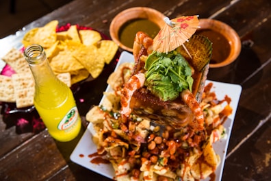 A vibrant Mexican dish featuring a large serving of shrimp cocktail topped with avocado and cilantro, garnished with a small paper umbrella. Surrounded by tortilla chips and crackers on a platter, accompanied by a bottle of lemon-lime flavored soda and bowls of green and red sauces on a wooden table.