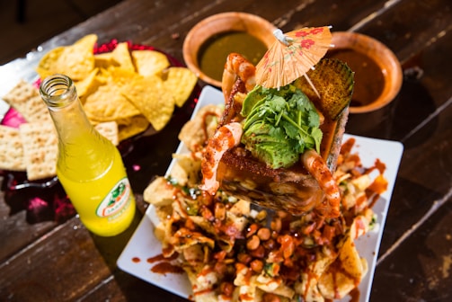 A vibrant Mexican dish featuring a large serving of shrimp cocktail topped with avocado and cilantro, garnished with a small paper umbrella. Surrounded by tortilla chips and crackers on a platter, accompanied by a bottle of lemon-lime flavored soda and bowls of green and red sauces on a wooden table.