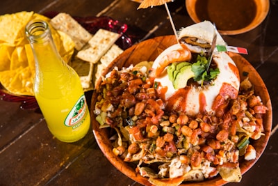 A colorful and vibrant dish is arranged on a wooden table. The dish appears to be a mix of various ingredients including nachos, beans, guacamole, and a drizzle of hot sauce, garnished with a slice of avocado and a Mexican flag toothpick. Nearby, there is an open bottle of Jarritos soda in pineapple flavor. Accompanying the meal are crackers and tortilla chips served in a red dish.