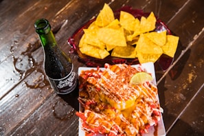 A glass bottle of soda sits on a wooden table beside a plate of nachos with a cheese dip topped with lime, chili powder, and drizzled sauce. A separate plate holds crispy tortilla chips.