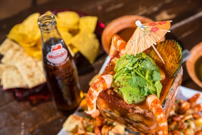 A vibrant and colorful seafood cocktail is topped with fresh avocado slices, cilantro, and surrounded by plump shrimp. An ornate paper umbrella adds a festive touch. Next to the dish, a glass bottle of soda is present, accompanied by crispy tortilla chips and slices of bread on a wooden table.