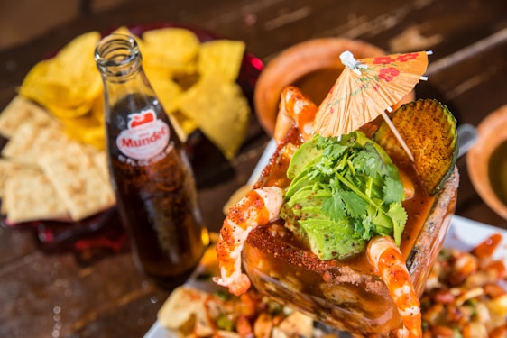 A vibrant and colorful seafood cocktail is topped with fresh avocado slices, cilantro, and surrounded by plump shrimp. An ornate paper umbrella adds a festive touch. Next to the dish, a glass bottle of soda is present, accompanied by crispy tortilla chips and slices of bread on a wooden table.
