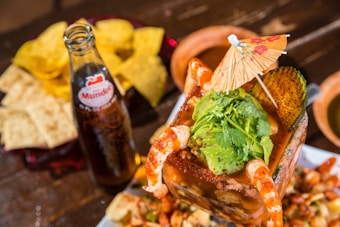A vibrant and colorful seafood cocktail is topped with fresh avocado slices, cilantro, and surrounded by plump shrimp. An ornate paper umbrella adds a festive touch. Next to the dish, a glass bottle of soda is present, accompanied by crispy tortilla chips and slices of bread on a wooden table.