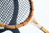 A vintage wooden tennis racket with a light brown frame and a tightly woven black string net. The frame features red accents with writing that includes the word 'Baumgarten' and a graphic at the intersection of the handle. The racket is casting a shadow on a white surface.
