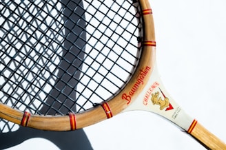 A vintage wooden tennis racket with a light brown frame and a tightly woven black string net. The frame features red accents with writing that includes the word 'Baumgarten' and a graphic at the intersection of the handle. The racket is casting a shadow on a white surface.