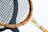 A vintage wooden tennis racket with a light brown frame and a tightly woven black string net. The frame features red accents with writing that includes the word 'Baumgarten' and a graphic at the intersection of the handle. The racket is casting a shadow on a white surface.