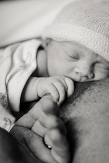 A newborn baby is sleeping peacefully, clutching an adult's finger. The baby is wearing a soft knit cap and is wrapped in a blanket. The image is in black and white, highlighting the delicate features of the baby and the gentle touch of the hand.