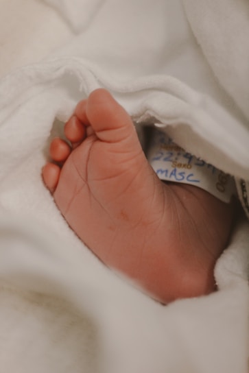 A close-up of a small, newborn baby's foot adorned with a hospital identification band, wrapped in soft, white fabric.