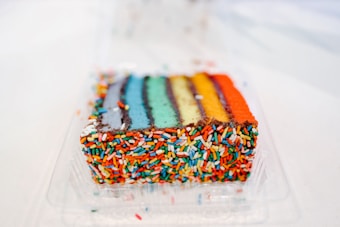 A slice of rainbow cake with multiple colored layers and covered in vibrant, multicolored sprinkles, placed inside a clear plastic container.