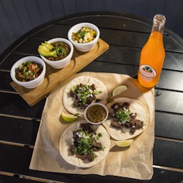 Three tacos filled with grilled meat, garnished with chopped onions and cilantro, placed on parchment paper on a dark table. A small cup of green salsa and lime wedges accompany the tacos. To the side, a glass bottle of Jarritos Mandarin soda stands next to the ensemble. Three small white bowls are placed on a wooden board, each containing different items: one with sliced avocado and sauce, the second with diced onions and corn, and the third with a fresh tomato salsa.