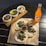 Three tacos filled with grilled meat, garnished with chopped onions and cilantro, placed on parchment paper on a dark table. A small cup of green salsa and lime wedges accompany the tacos. To the side, a glass bottle of Jarritos Mandarin soda stands next to the ensemble. Three small white bowls are placed on a wooden board, each containing different items: one with sliced avocado and sauce, the second with diced onions and corn, and the third with a fresh tomato salsa.