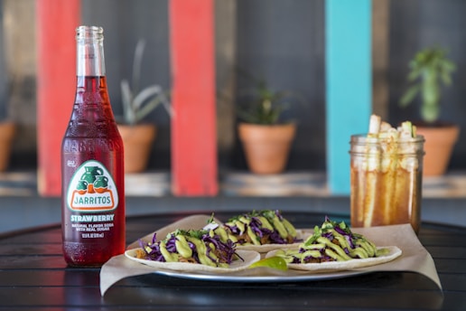 A bottle of Jarritos strawberry soda sits beside a plate of tacos with vibrant toppings and a jar of snack sticks, all set on a dark surface. The background features potted plants against a backdrop with colorful wooden panels.