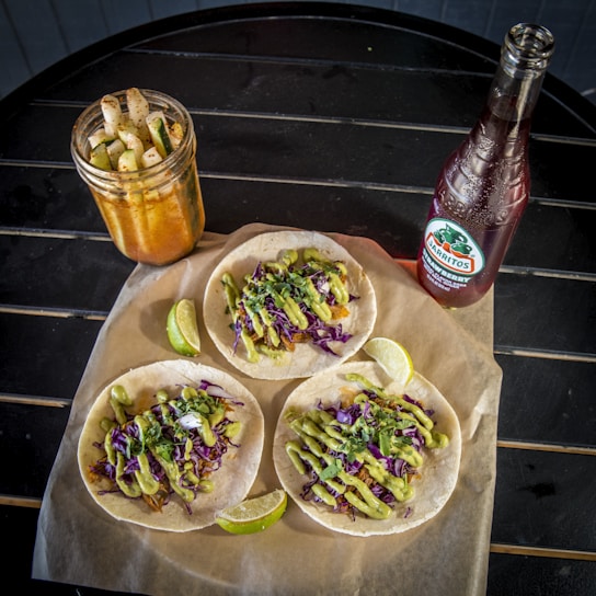 Three tacos are arranged on a piece of brown parchment paper atop a round black table. The tacos are topped with avocado sauce, shredded purple cabbage, and green herbs. Beside the tacos, there are lime wedges. To the left, a mason jar filled with pickled vegetables, including cucumber and jicama, is visible. On the right, there's a bottle of Jarritos soda.